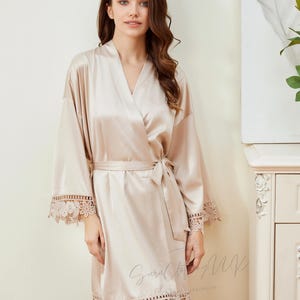 May include: A light beige satin robe with lace trim around the sleeves and hem. The robe has a tie belt and a V-neck.