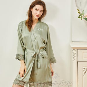 May include: A sage green satin robe with lace trim and a personalized name, "Karis", embroidered on the front. The robe has a tie belt and is shown on a model.