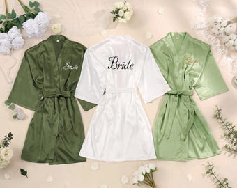 Puede incluir: Tres batas de satén con cinturón y mangas anchas. La bata de la izquierda es de color verde oliva oscuro con el nombre Sadie bordado en blanco. La central es blanca con la palabra Bride en negro. La de la derecha es verde salvia con bordado dorado.