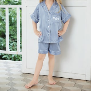 May include: A light blue satin pajama set with short sleeves and shorts. The top has a button-up front and a pocket on the left chest with the word "Babe" embroidered in white.