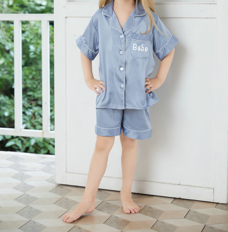 May include: A light blue satin pajama set with short sleeves and shorts. The top has a button-up front and a pocket on the left chest with the word "Babe" embroidered in white.