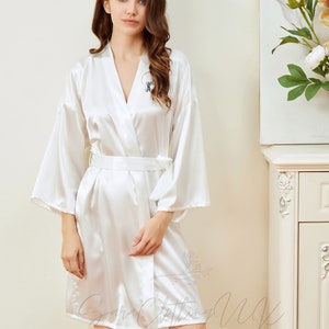 May include: White satin robe with a black embroidered initial "K" on the left side of the chest. The robe has long sleeves and a tie belt.