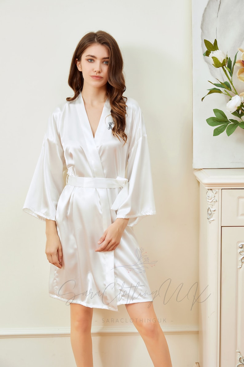 May include: White satin robe with a black embroidered initial "K" on the left side of the chest. The robe has long sleeves and a tie belt.