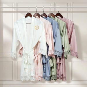 May include: A set of six satin robes with lace trim, hanging on hangers. The robes are in white, pink, blue, green, and blush. The white robe has a gold embroidered letter N on the front.