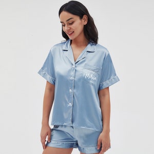 May include: A light blue satin pajama set with short sleeves and shorts. The top has a button-up front and a pocket on the left chest with the name "Abbie" embroidered in white.