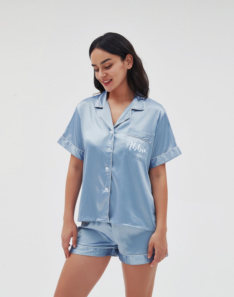 May include: A light blue satin pajama set with short sleeves and shorts. The top has a button-up front and a pocket on the left chest with the name "Abbie" embroidered in white.