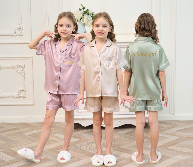 May include: Three girls wearing matching satin pajama sets. One set is light pink, one is beige, and the third is sage green. The sage green set has "Flower girl" printed on the back. Each set includes a short-sleeved button-up shirt and shorts.