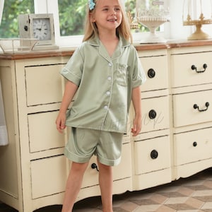 May include: A young girl wearing a light green satin pajama set with short sleeves and shorts. The set has white piping and a button-up front. The girl is standing in front of a white dresser with drawers.