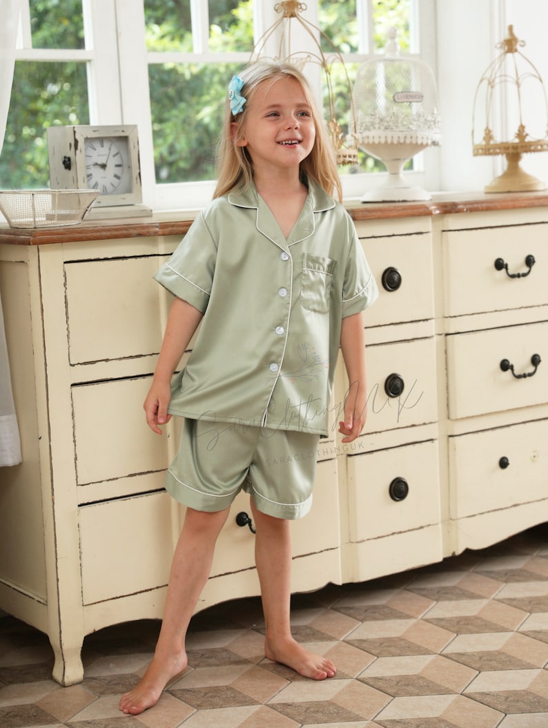 May include: A young girl wearing a light green satin pajama set with short sleeves and shorts. The set has white piping and a button-up front. The girl is standing in front of a white dresser with drawers.