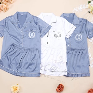 May include: Three sets of pajamas, one white and two blue. The blue pajamas have a white trim and a pocket with a white embroidered wreath and letter. The white pajamas have a black trim and the text "CMR" embroidered on the pocket.