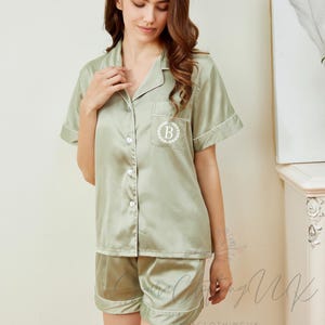 May include: Sage green pajama set with white trim. The short-sleeved top has a button-down front and a breast pocket with a monogram. The matching shorts have a similar white trim. The set is made of a silky material.