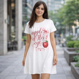 Puede incluir: Vestido camiseta blanco con diseño de rayas verticales rosas. La parte delantera presenta el texto "apple of His eye" en fuente cursiva, un lazo rosa y una ilustración de manzana roja. También se incluye el texto "Salmo 17:8".