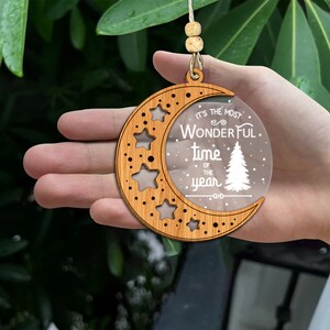 May include: Wooden Christmas ornament shaped like a crescent moon with a clear acrylic insert. The insert has the text "It's the most wonderful time of the year" with a stylized pine tree and snowflakes.