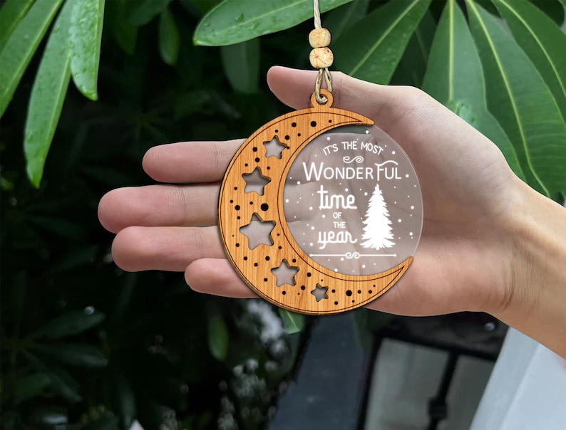 May include: Wooden Christmas ornament shaped like a crescent moon with a clear acrylic insert. The insert has the text "It's the most wonderful time of the year" with a stylized pine tree and snowflakes.
