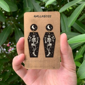 May include: A pair of black coffin-shaped earrings with a skeleton dancing inside and a crescent moon charm hanging above. The earrings are on a brown card with the word "WALLNEOSS" printed on it.