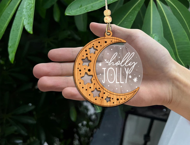 May include: A wooden crescent moon ornament with a clear acrylic insert. The insert has a white design that says "holly jolly" with snowflakes and stars.