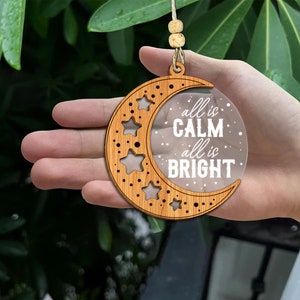 May include: Wooden crescent moon ornament with a clear acrylic insert that reads "all is calm, all is bright". The ornament is decorated with stars.