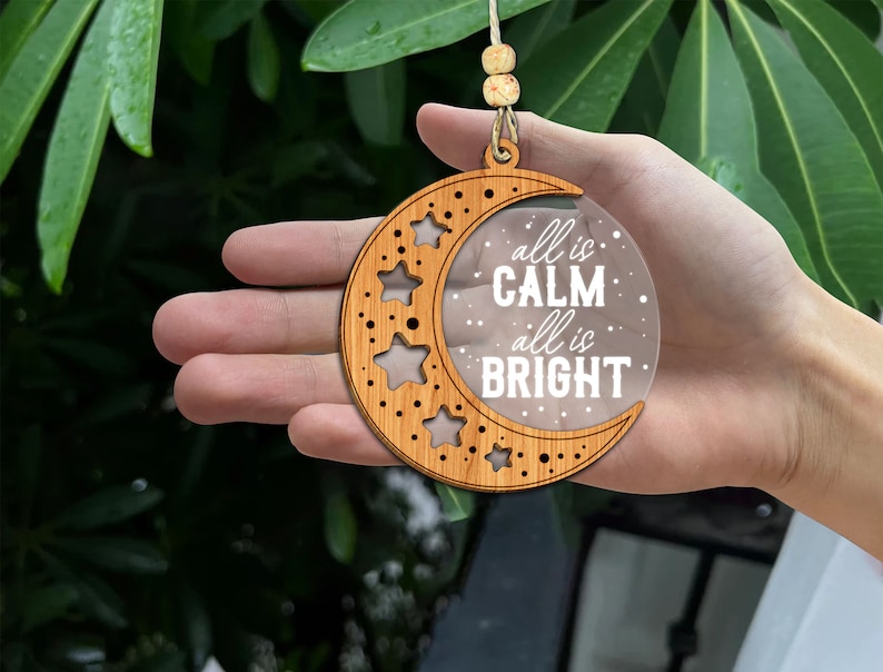 May include: Wooden crescent moon ornament with a clear acrylic insert that reads "all is calm, all is bright". The ornament is decorated with stars.