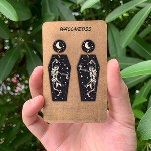 May include: A pair of black coffin-shaped earrings with a white skeleton dancing inside and a crescent moon charm hanging from the top. The earrings are displayed on a brown card with the text "WALLNEOSS" printed on it.
