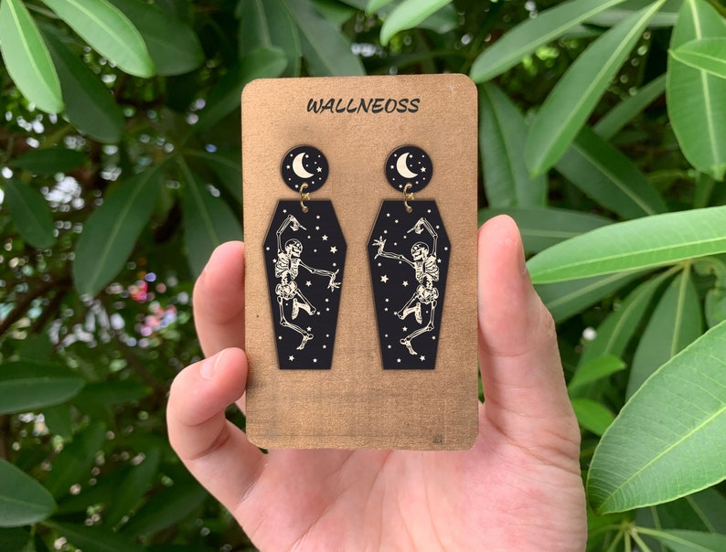May include: A pair of black coffin-shaped earrings with a white skeleton dancing inside and a crescent moon charm hanging from the top. The earrings are displayed on a brown card with the text "WALLNEOSS" printed on it.