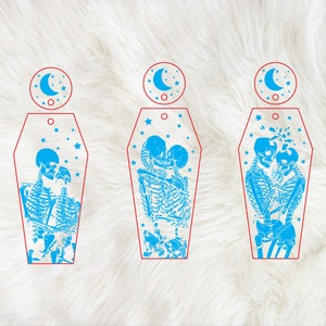 Op de afbeelding: Drie blauwe en witte kistvormige ornamenten met een skeletpaar dat elkaar kust in elk ornament. Elk ornament heeft een halve maan en sterren op de achtergrond.