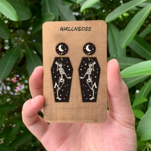 May include: A pair of black coffin-shaped earrings with a skeleton dancing inside and a crescent moon charm hanging from the top. The earrings are on a card that says "WALLNEOSS".