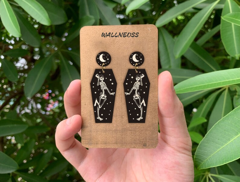 May include: A pair of black coffin-shaped earrings with a skeleton dancing inside and a crescent moon charm hanging from the top. The earrings are on a card that says "WALLNEOSS".