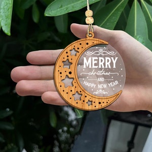 May include: A wooden crescent moon ornament with a clear acrylic insert that reads "Merry Christmas and Happy New Year". The ornament is decorated with stars and has a hole at the top for hanging.