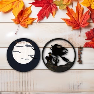 Op de afbeelding: Twee ronde houten Halloween-decoraties. E&eacute;n toont een maan, vleermuizen en de tekst "Wishing You a Happy". De andere toont een heks op een bezem die langs een spookhuis vliegt. Herfstbladeren zijn verspreid.