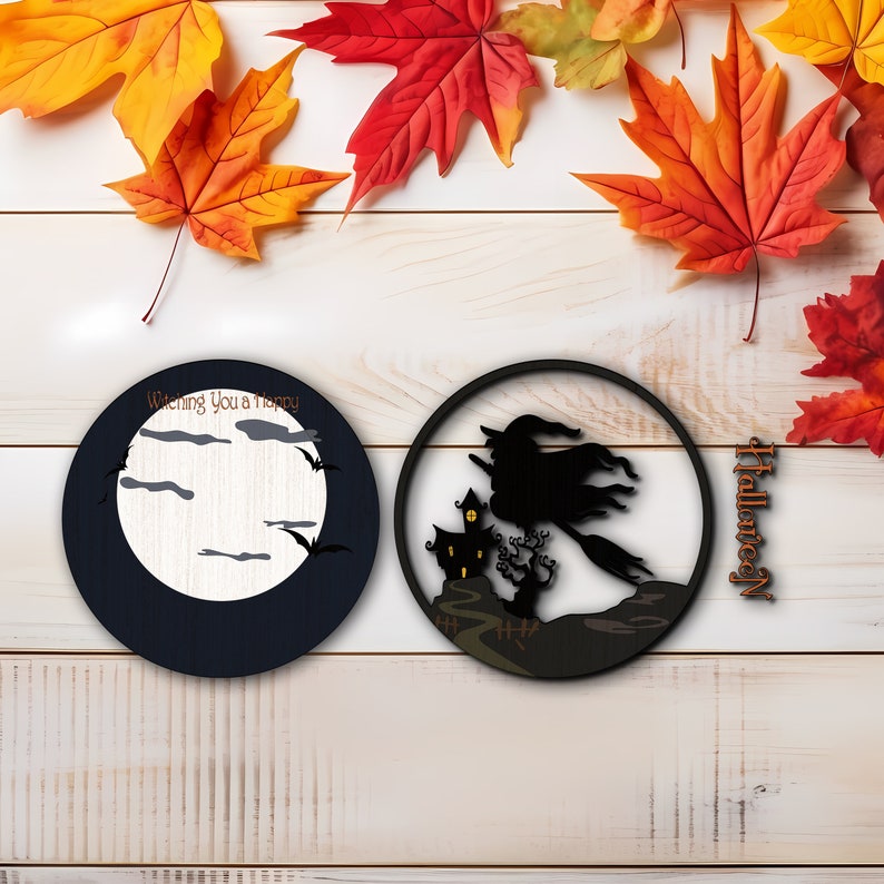 Op de afbeelding: Twee ronde houten Halloween-decoraties. E&eacute;n toont een maan, vleermuizen en de tekst "Wishing You a Happy". De andere toont een heks op een bezem die langs een spookhuis vliegt. Herfstbladeren zijn verspreid.