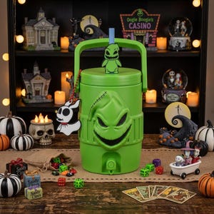 May include: A bright green beverage cooler featuring a molded Oogie Boogie face on the front, a top handle, and a blue straw. A small Oogie Boogie figure sits on the lid, and a white Zero character charm hangs from a red beaded chain on the side.