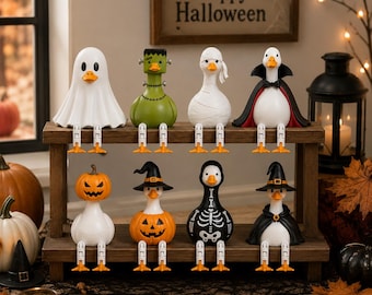 May include: Eight decorative duck figurines arranged on a two-tier wooden shelf, each dressed in different Halloween costumes including a ghost, Frankenstein, mummy, vampire, pumpkin head, witch, and skeleton, all featuring dangling orange feet.