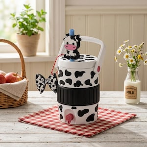 May include: A white beverage dispenser with a black cow print pattern, featuring a black silicone grip band, a red spigot, a white handle, and a blue straw topped with a black and white cow figure. A black and white bow charm hangs from the straw.