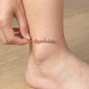 Op de afbeelding: Roségouden enkelband met het woord "dimpledbabe" in cursief schrift. De delicate ketting wordt bij de enkel vastgehouden. De achtergrond is een lichtbruin houten oppervlak.