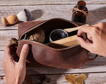 May include: A brown leather toiletry bag with a zipper closure. The bag is open and contains a wooden comb, a small camera, and a ball of twine.