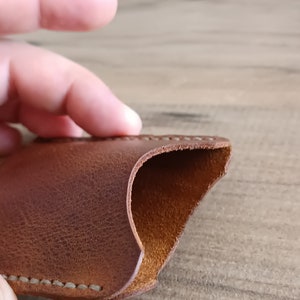 May include: Close-up of a brown leather case with visible stitching along the edges. The case has a curved opening, revealing a lighter brown interior. The leather has a textured appearance.