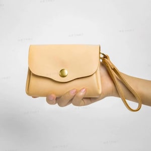 May include: A small, beige leather wallet with a flap closure and a gold-colored button. The wallet has a wrist strap and is held in a hand. The leather has a natural, unpainted appearance.