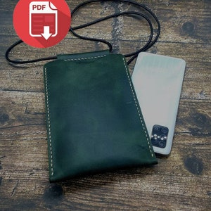 Può includere: Una custodia per telefono in pelle verde con dettagli di cucitura bianca e un cinturino nero. La custodia è appoggiata su una superficie di legno con uno smartphone accanto.