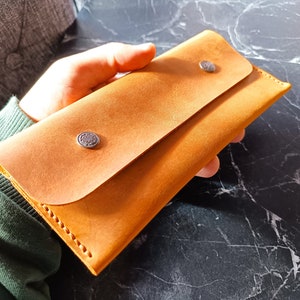 May include: A tan leather wallet with a flap closure and two silver snaps. The wallet is held in a hand.