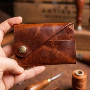May include: A brown leather wallet with a button closure and a diagonal design. The wallet has two card slots and is held in a hand. The leather has a distressed look.