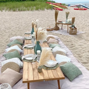 May include: A beach picnic setup with a wooden table, adorned with plates, candles, and decorative vases. Pillows in shades of green, beige, and white are arranged on a blanket. Pampas grass and other natural elements add to the aesthetic.