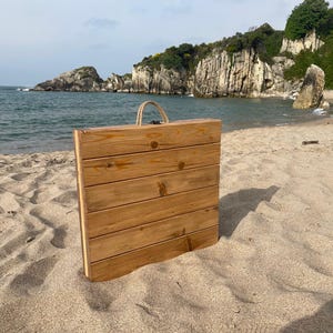 May include: A wooden folding table with a rope handle stands on a sandy beach. The table is made of light brown wooden planks. The background features a scenic view of the ocean, cliffs, and a cloudy sky.