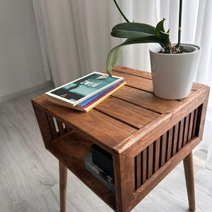 May include: A brown wooden side table with a slatted design and a lower shelf. A stack of books and a potted orchid plant are on top. The book cover reads "ZWEIG". The table is likely made of solid wood.