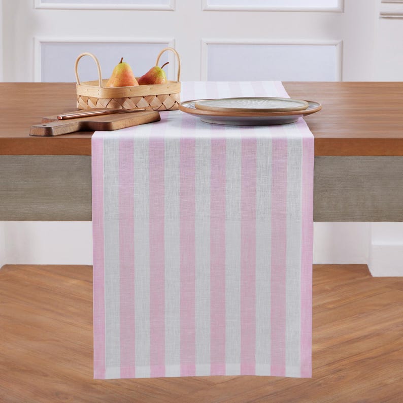 May include: A pink and white striped table runner is draped over a wooden table. A woven basket with pears, a wooden cutting board, and a plate are on the table. The runner adds a decorative touch to the dining area.