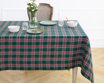 May include: A rectangular tablecloth featuring a dark green, red, and white plaid pattern. The table is set with two white bowls, two green plates, two clear glasses, gold-toned cutlery, and a glass vase containing white flowers.