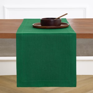 May include: A solid emerald green rectangular table runner with a stitched border detail. The runner is draped over a wooden table and topped with a dark brown wooden bowl, a matching wooden plate, and a wooden spoon resting inside the bowl.