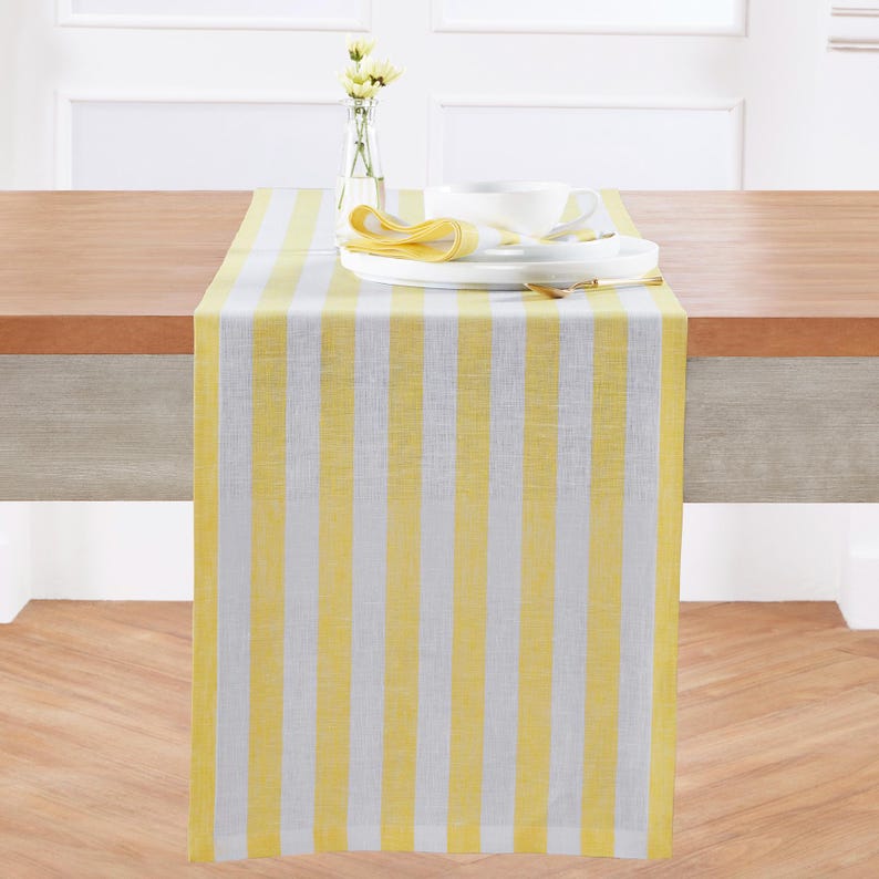 May include: A yellow and white striped table runner is draped across a wooden table. White plates, a cup, and a yellow napkin are on top of the runner. A small vase with yellow flowers is in the background.