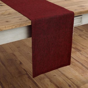 May include: A rectangular table runner in a deep burgundy red color with a subtle, dark, mottled texture throughout the fabric. The runner is draped over the edge of a wooden table, showing a clean, straight hem along the bottom edge.