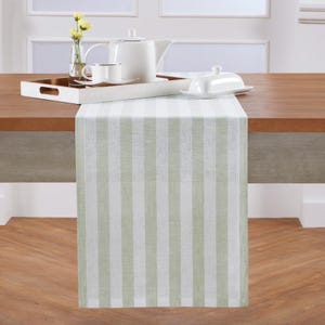 May include: A green and white striped table runner. The runner is made of a light-colored fabric and is draped over a wooden table. A white tea set and a tray with a gold rim are on the table.