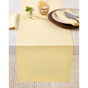 May include: A pale yellow table runner with a subtle border detail, set on a light-colored table. The runner is adorned with a place setting, a glass of water with a citrus slice, and a small vase of flowers. The scene suggests a dining or event setting.
